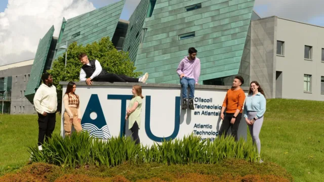 Students sitting and standing around the ATU sign on the Galway City campus, with the iconic green sails building in the background on Dublin Road, Galway.