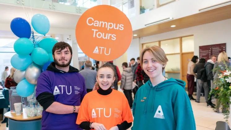 Three student ambassadors wearing ATU branded jumpers and t-shirts. One student is wearing a purple ATU jumper and holding up an orange sign that reads 'Campus Tours.' All are smiling at the camera.