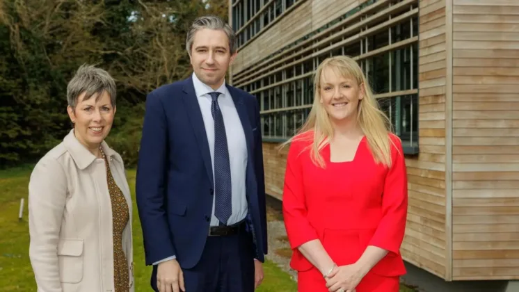 Three people standing outside a wooden-sheeted building, with the man in the centre and two women either side. All are smiling and looking at the camera.