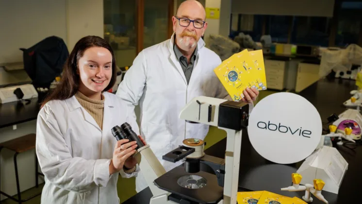 Oli Melia, ATU Event Manager and Science Festival Producer, stands in a lab holding up flyers for the Science Festival, while Katie Aherne, NPI Process Engineer at AbbVie, looks through a microscope.