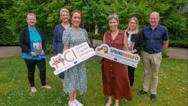 A group of six people standing in a leafy environment with feet shoulder-width apart, promoting the Springboard initiative. The two women at the front are holding signs, while two people on each side are holding books. The rest of the group stands behind them, all smiling