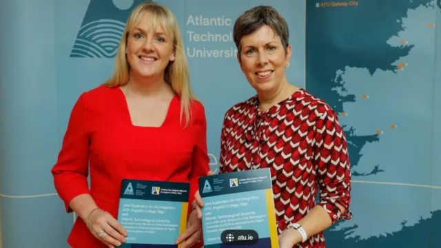 Two women standing together, both holding an application form, smiling and looking at the camera