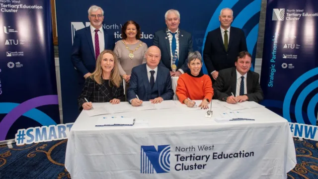 Four people seated at a table, signing a document with pens in their hands. Behind them, four people are standing. The tablecloth reads 'North West Tertiary Education Cluster.