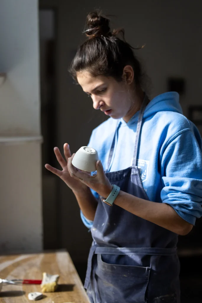 Female student wearing apron molding clay in ceramics workshop. 