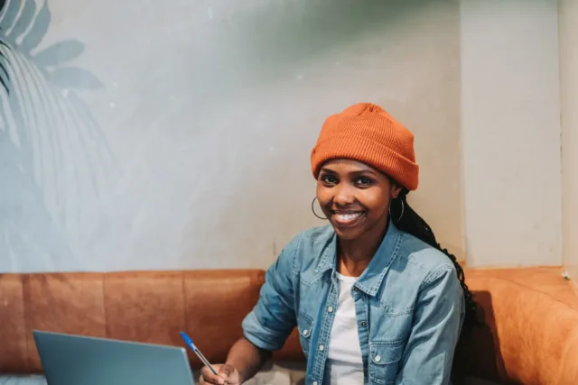 Female international student sits at table in cafe with laptop, wearing denim shirt and orange beanie hat