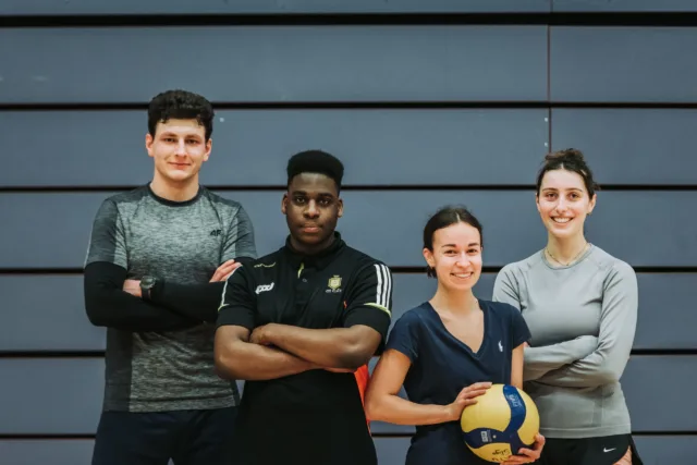 Four students, two boys and two girls, stand in sports gear with arms folder. One girl holding ball.