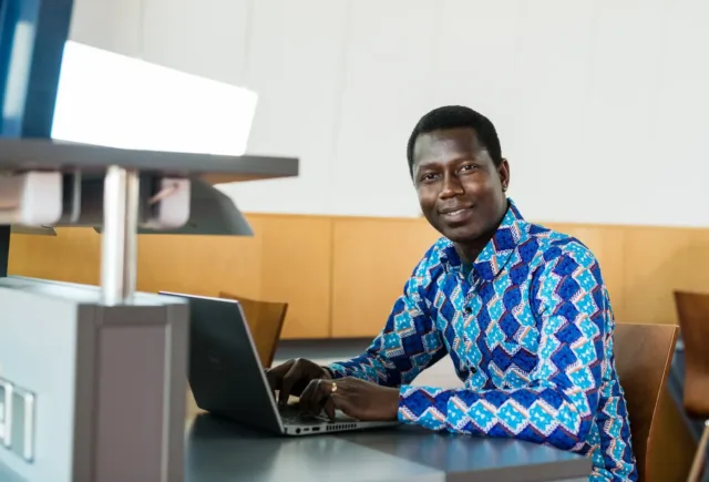 Male international student sits at desk in library typing on laptop, wearing a blue patterned shirt