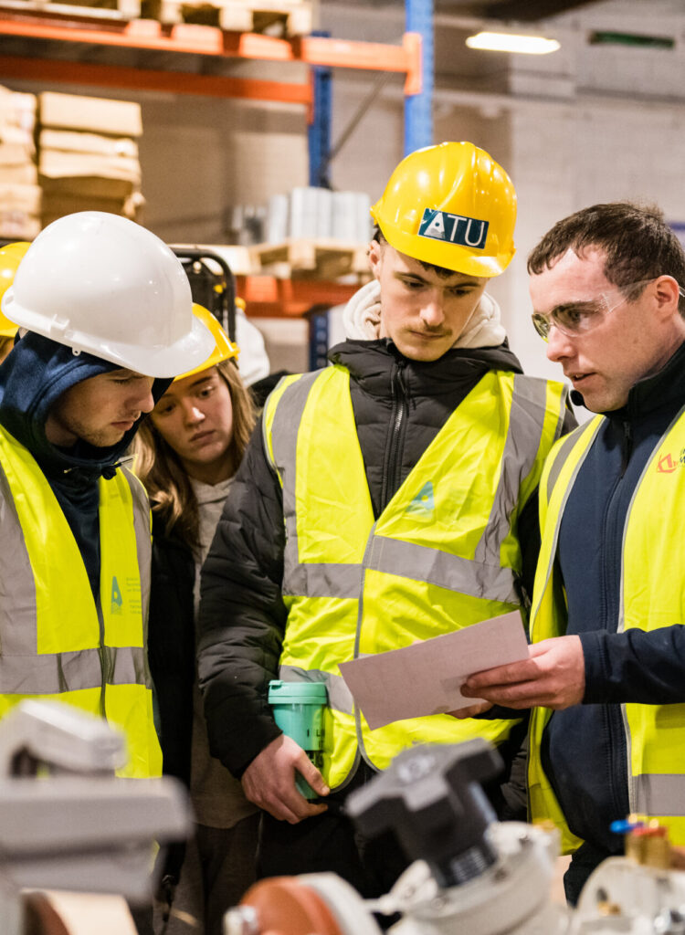 Students in ATU branded hard hats and high visibility jackets look at engineering plans in a warehouse