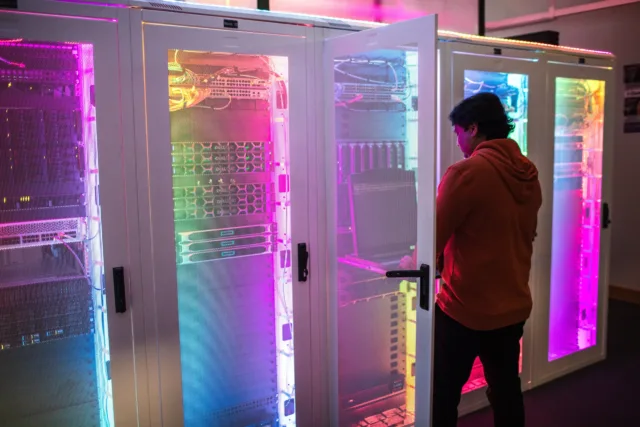 Male international student stands in front of computer servers lit up by multi-colour LED lighting