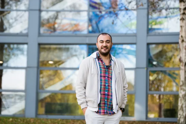 Male international student stands with hands in pocket in front of campus building