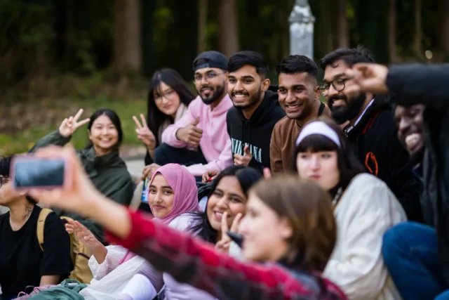 A group of international students sitting on steps outside the campus, smiling while taking a group selfie photo.