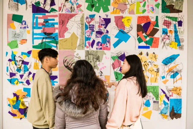 Three international students, one boy and two girls, looking at a collage wall of colour art and paper.