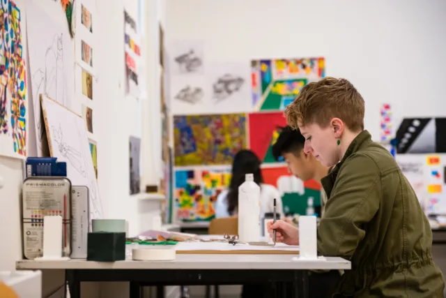 Student works at desk on artwork, surrounded by artwork on surrounding walls