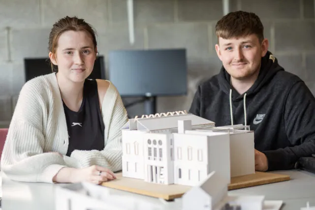 Male and Female student sit in computer lab with model home on desk