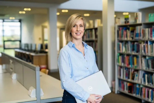 Female mature student standing in the library holding a laptop