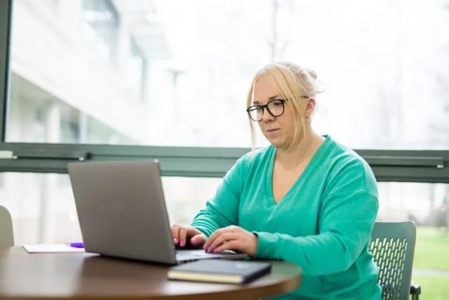 Female student in green jumper sits by window working on a laptop