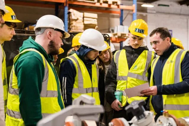 Engineering students in hard-hats and high visibility vest stand in warehouse looking at plans