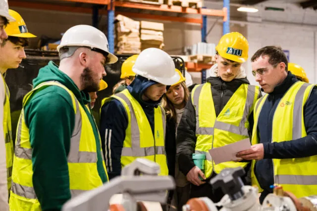 Engineering students in hard-hats and high visibility vest stand in warehouse looking at plans